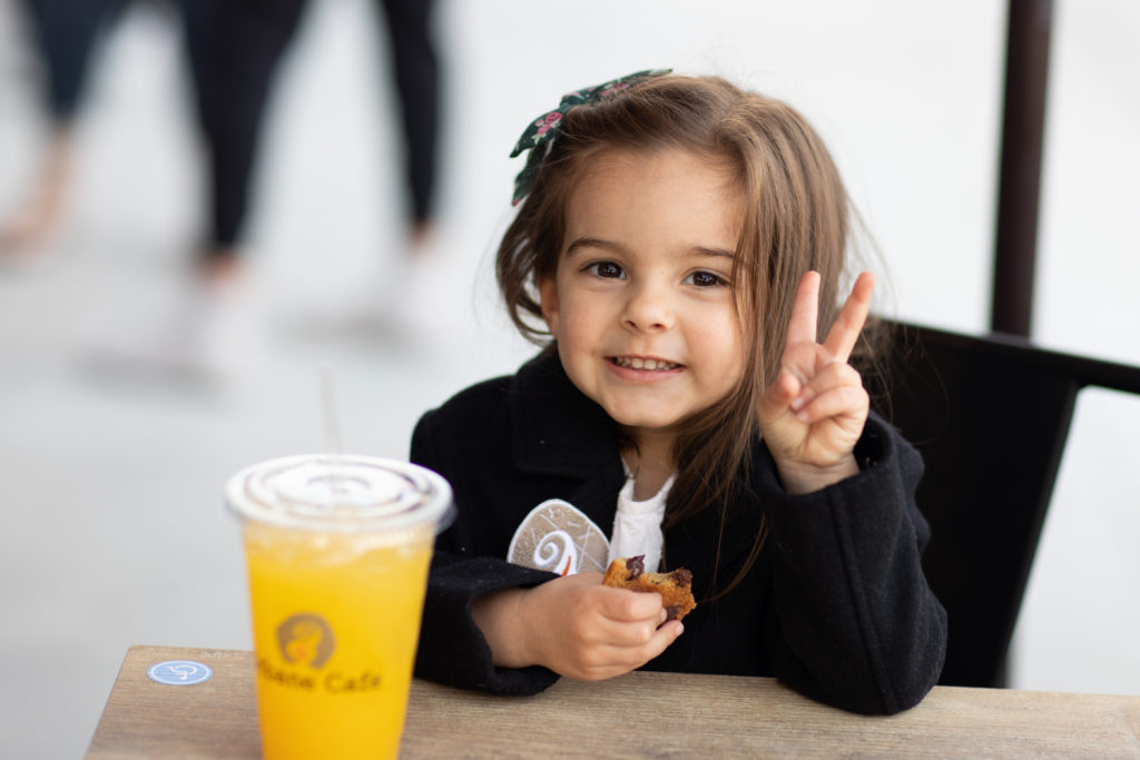 little girl smiling holding up peace sign holding cookie with urbane cafe drink